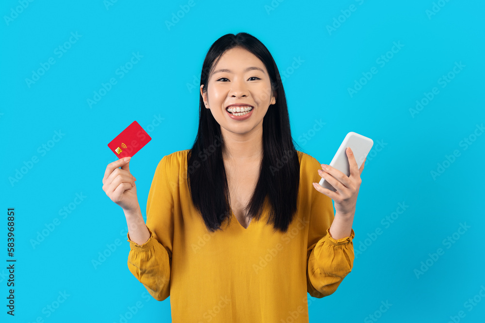 Happy asian woman holding smartphone and bank card on blue Happy asian woman holding smartphone and bank card on blue