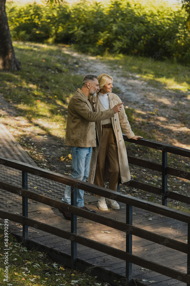 Fototapeta premium Smiling mature man pointing with finger near blonde wife while standing on bridge in park.