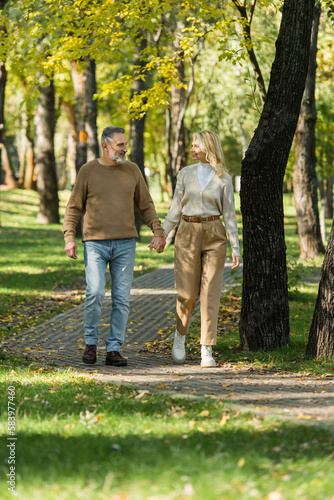 full length of stylish middle aged couple holding hands and smiling while walking together in park during springtime.