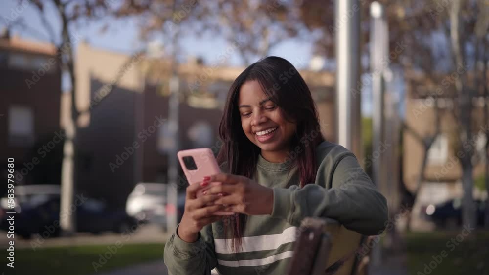 Teen Latin girl looks at cell phone smiling funny. Happy young woman sitting in the park using her mobile. People cheerful checking their social networks outside. Concept of online relationships. Teen Latin girl looks at cell phone smiling funny. Happy young woman sitting in the park using her mobile. People cheerful checking their social networks outside. Concept of online relationships.