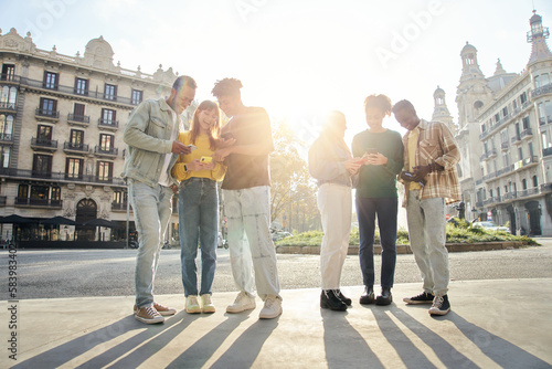 Group of smiling multiracial young people using cell phones. Cheerful students standing with technological devices. Happy university classmates on school trip in a European city. Persons outdoors photo