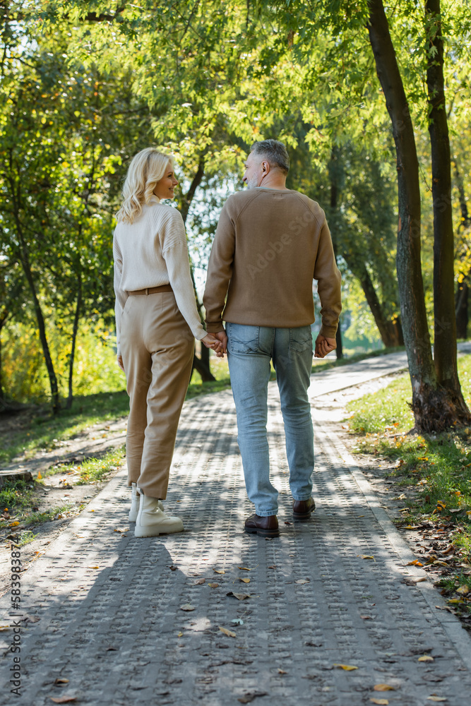 full length of stylish middle aged couple holding hands while walking together in green park. full length of stylish middle aged couple holding hands while walking together in green park.