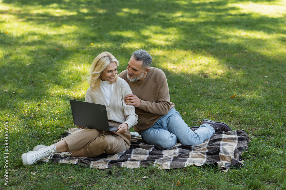 carefree middle aged husband and wife watching movie on laptop and sitting on blanket in park. carefree middle aged husband and wife watching movie on laptop and sitting on blanket in park.