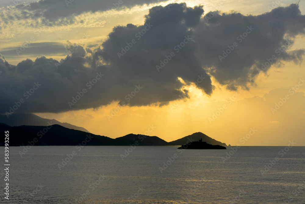 Sea sunset behind the clouds. In the distance are mountains in shadow and an island with a small lighthouse