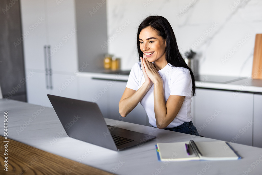 Young woman looking her laptop while holding a cup of coffee in the kitchen at home. Young woman looking her laptop while holding a cup of coffee in the kitchen at home.