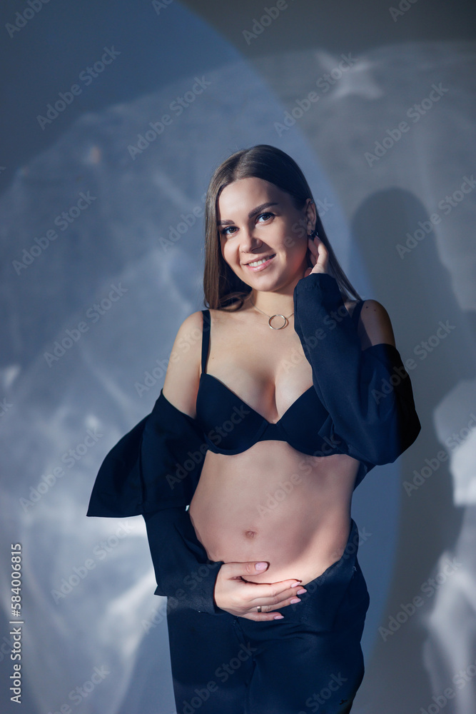 Obraz premium Portrait of a young pregnant woman in a black classic suit. A pregnant woman on a gray background hugs her belly.
