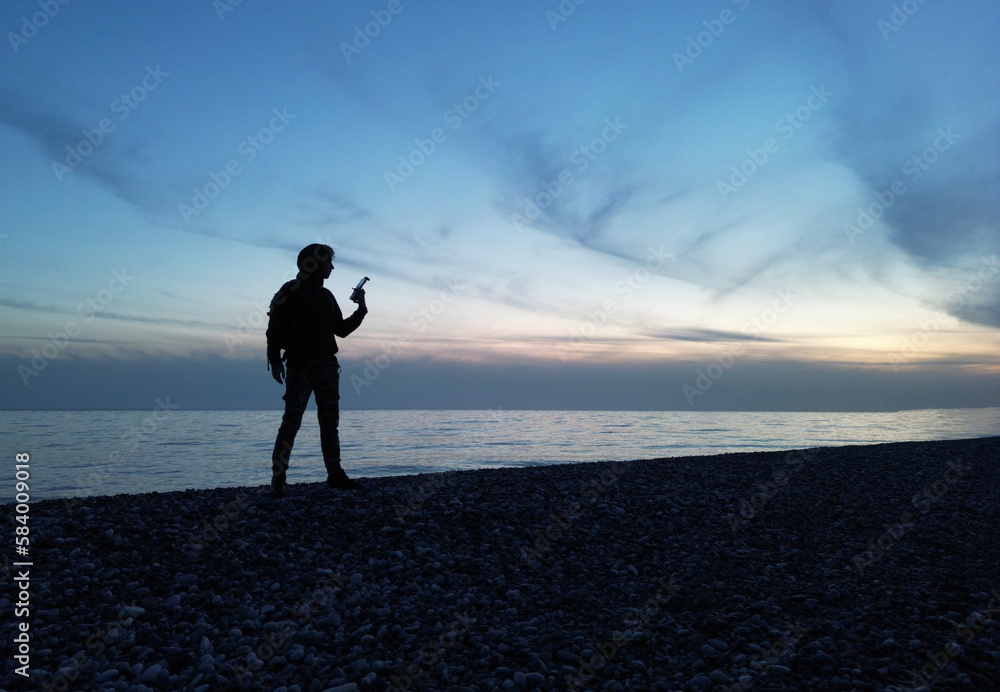 the silhouette of a guy at a beautiful sunset against the background of the sea and clouds in his hand is a remote control from a drone