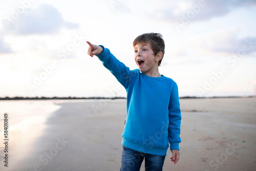 Surprised child stretched out his hand forward, pointing his finger, sea and the sky with clouds in background. Beach, sunset light. Concept of travel, vacation, childhood. Caucasian boy 6 years old.