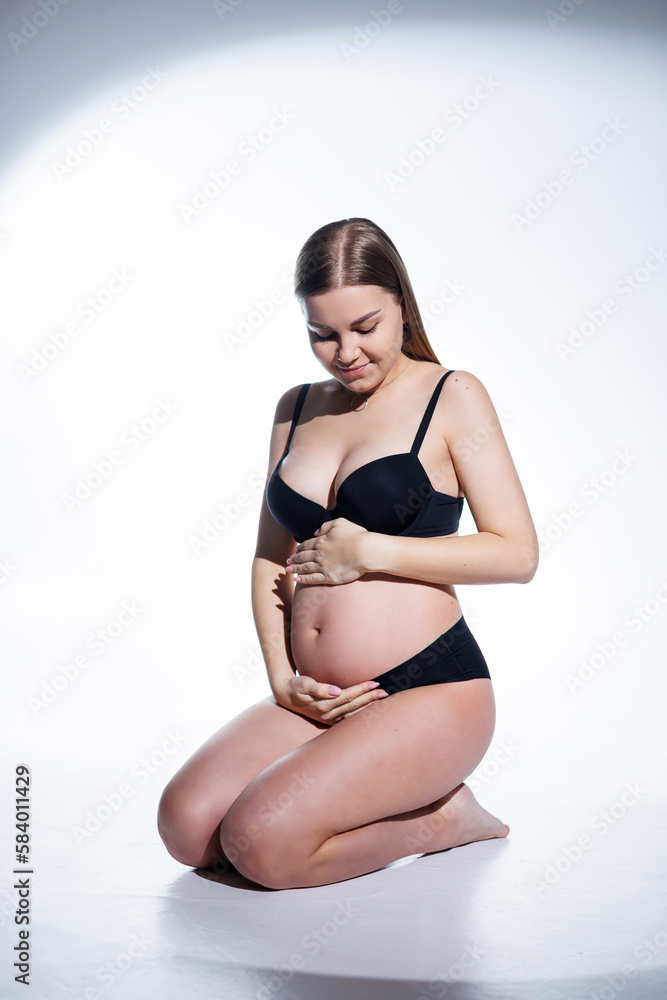 Obraz premium Cute pregnant woman with long hair in black underwear is sitting on a white background. Women's underwear for pregnant women.