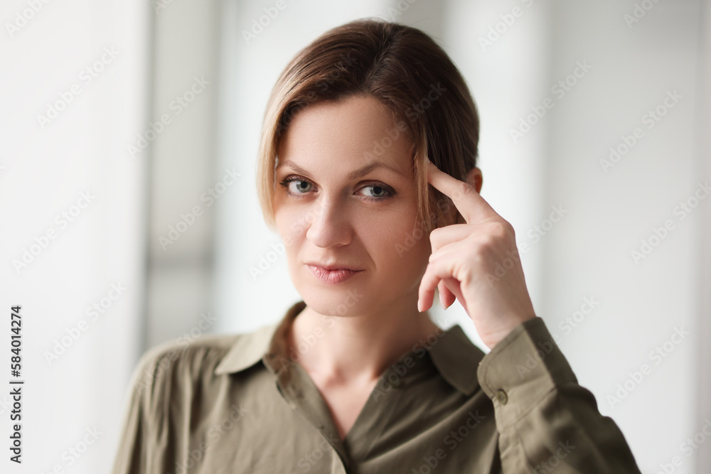 Serious woman puts index finger on temple looking in camera Serious woman puts index finger on temple looking in camera