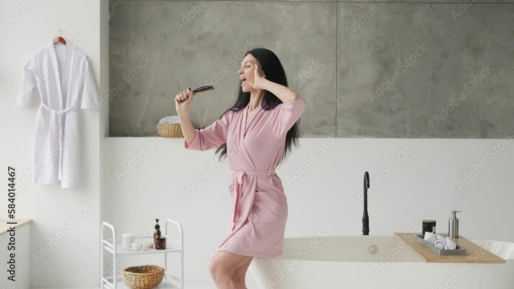 Cheerful Brunette woman Singing With Hairbrush Having Fun In Modern Bathroom Indoor. Haircare Cosmetics Advertisement. Beauty Rituals Concept Cheerful Brunette woman Singing With Hairbrush Having Fun In Modern Bathroom Indoor. Haircare Cosmetics Advertisement. Beauty Rituals Concept