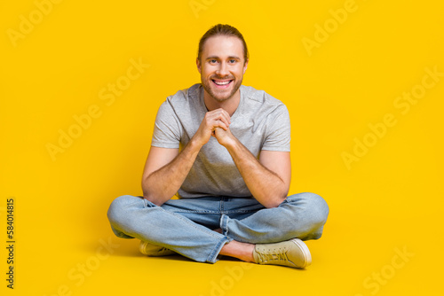 Full length photo of pleasant handsome guy wear light t-shirt jeans sneakers outfit sitting on floor isolated on yellow color background © deagreez