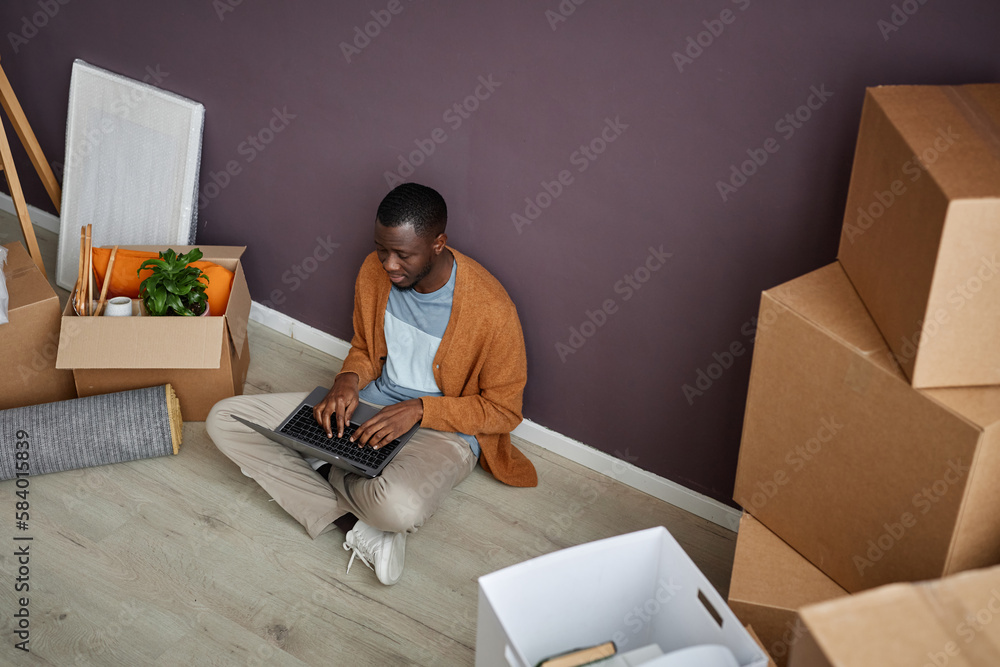 Fototapeta premium African American man sitting on the floor among boxes and typing on laptop during relocation