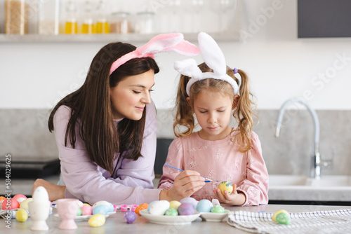 Young mother painting Eater eggs with her child girl daughter 4-5 year old at kitchen table together. Holiday season celebration. Springtime. Motherhood.