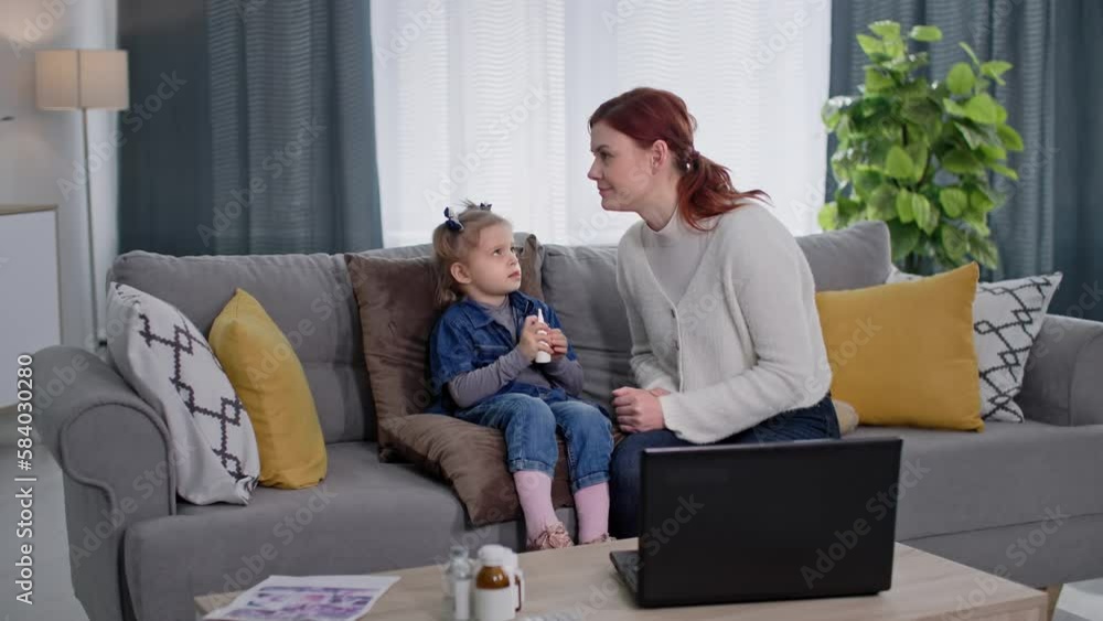 caring female parent is persuading a little girl to spray us with a runny nose spray while sitting on couch in room, child is looking at camera caring female parent is persuading a little girl to spray us with a runny nose spray while sitting on couch in room, child is looking at camera