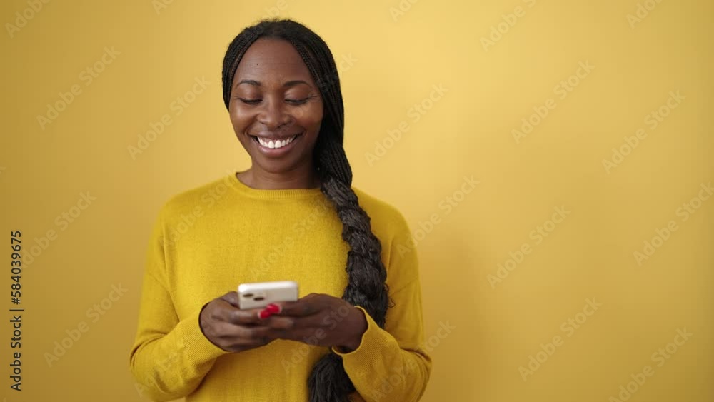 African woman smiling using smartphone over isolated yellow background African woman smiling using smartphone over isolated yellow background
