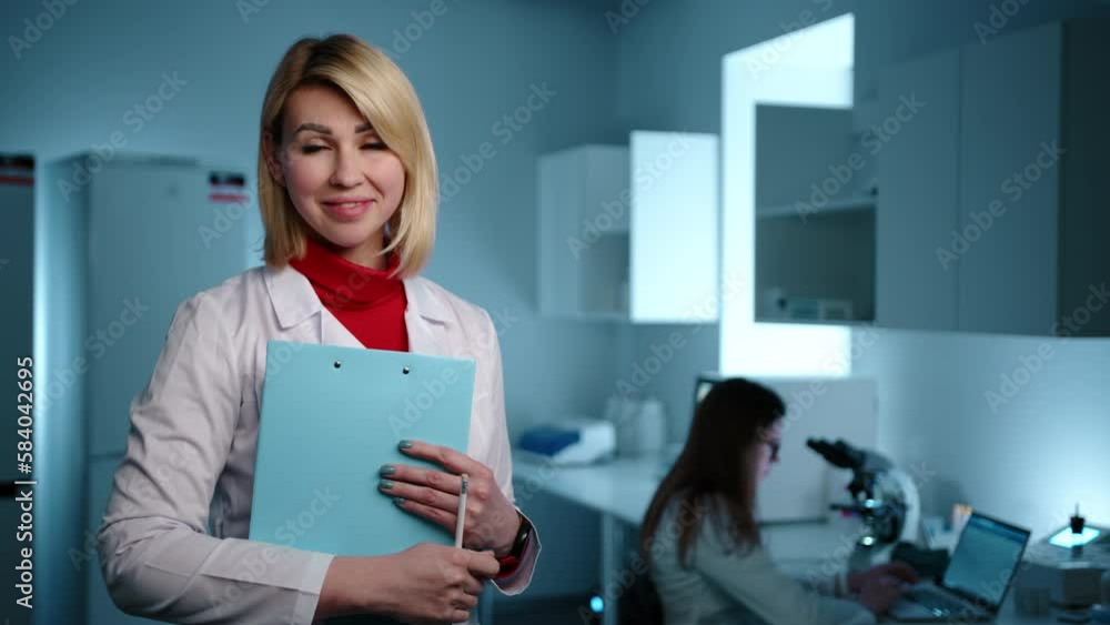 A blonde female doctor in a laboratory writing something on a tablet and then smiling at the camera. Another doctor is working on a microscope in the background. High quality 4k footage A blonde female doctor in a laboratory writing something on a tablet and then smiling at the camera. Another doctor is working on a microscope in the background. High quality 4k footage