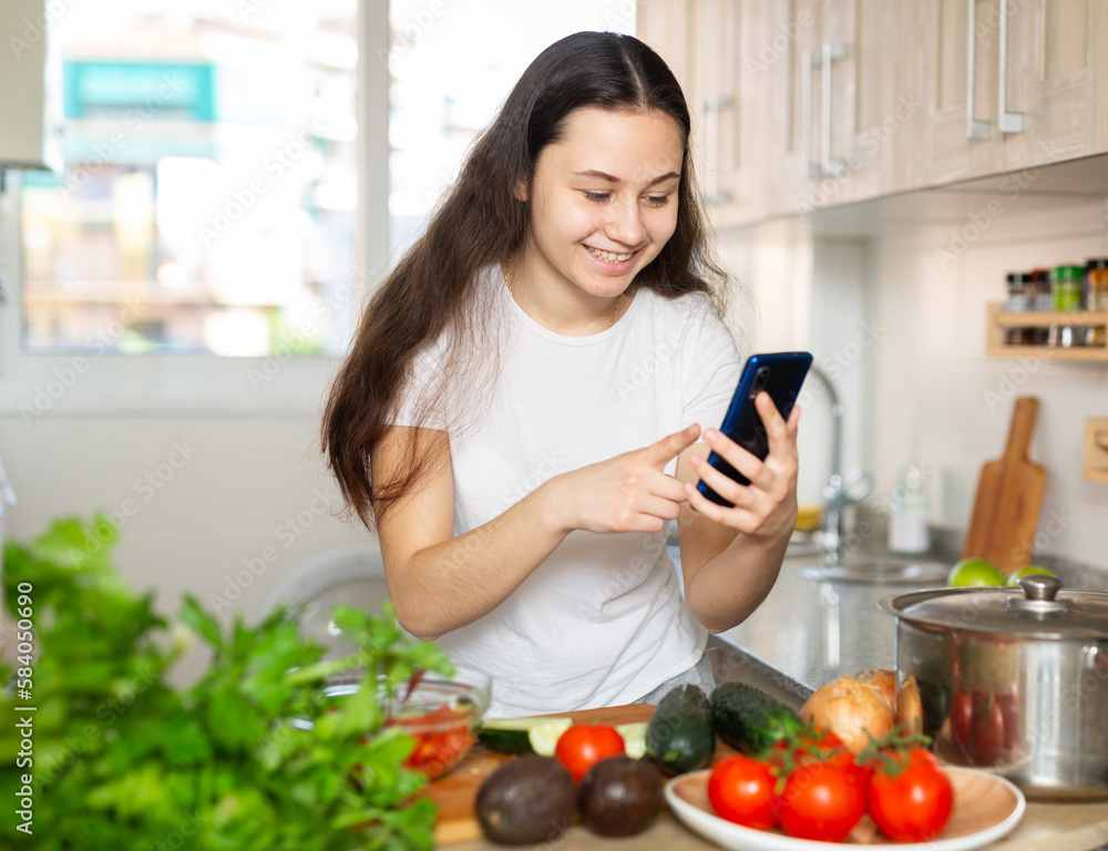 Portrait of attractive pretty girl reading recipe online on the phone in the kitchen