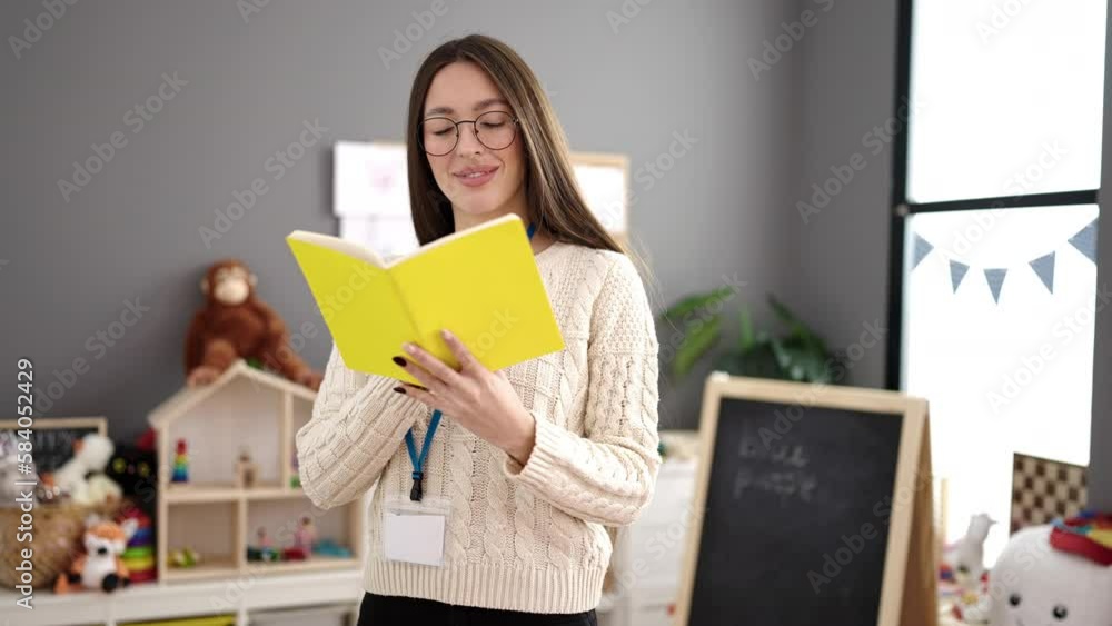 Young beautiful hispanic woman preschool teacher reading book standing at kindergarten Young beautiful hispanic woman preschool teacher reading book standing at kindergarten