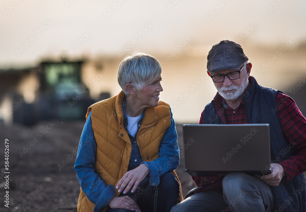 Senior farmers working on laptop in field with tractor in background