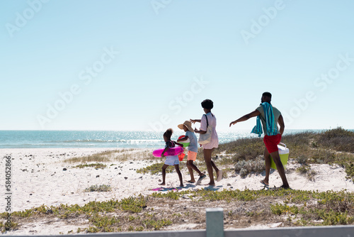 African american parents and children with cooler, swimming float, ball walking at beach under sky