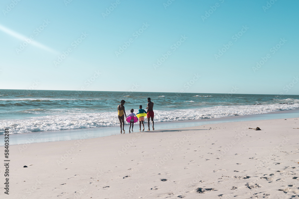 Rear view of african american parents and children with swimming float running towards sea under sky Rear view of african american parents and children with swimming float running towards sea under sky