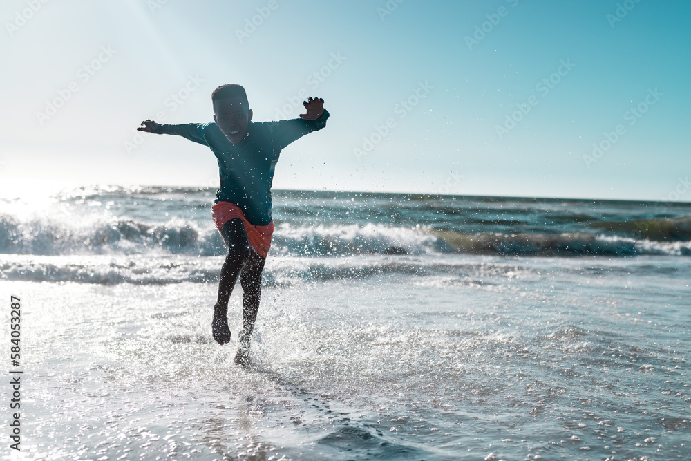Full length of carefree african american boy running and splashing water in sea against clear sky