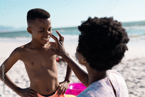 African american mother applying sunscreen on son's nose at beach against clear sky, copy space