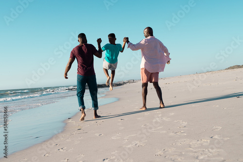 Rear view of african american father and grandfather holding boy's hands and picking him up at beach