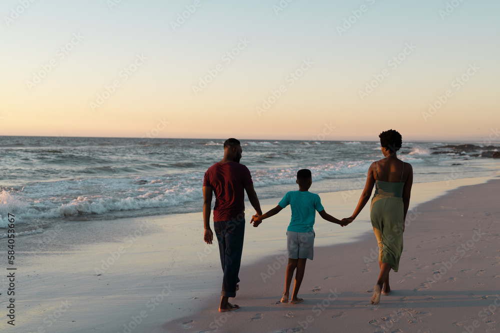 Rear view of african american boy holding mother and father's hands and walking on beach at sunset