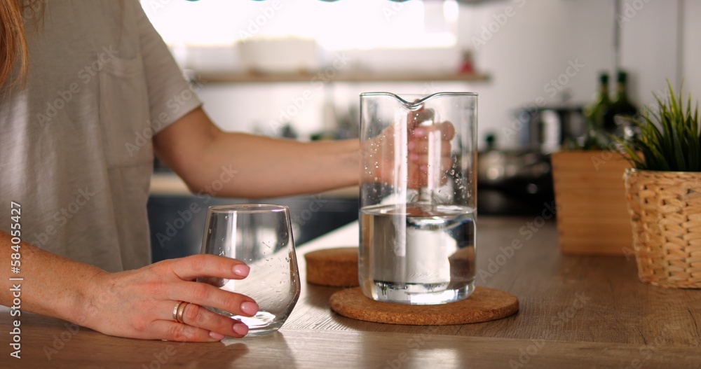 Unrecognizable young girl or woman holds glass jar water in kitchen. Hand of anonymous person in white shirt take jug from countertop