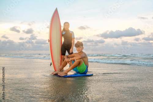 Teenage boy and girl sitting and standing with surfboard and boogey board on beach wearing swimwear during sunset photo