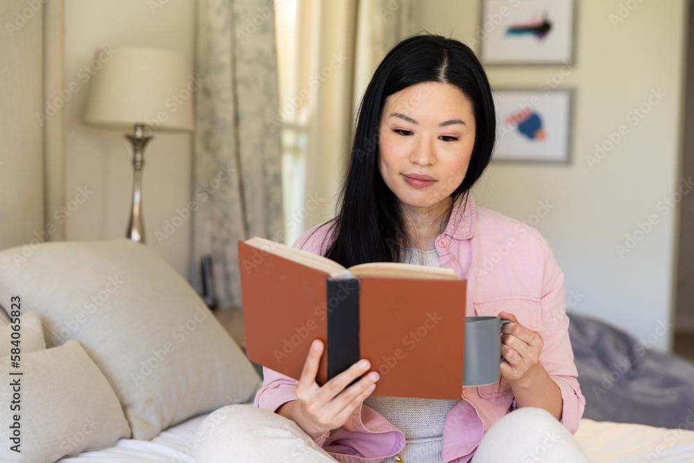 Smiling asian woman sitting on her bed reading a book and drinking coffee during the day Smiling asian woman sitting on her bed reading a book and drinking coffee during the day