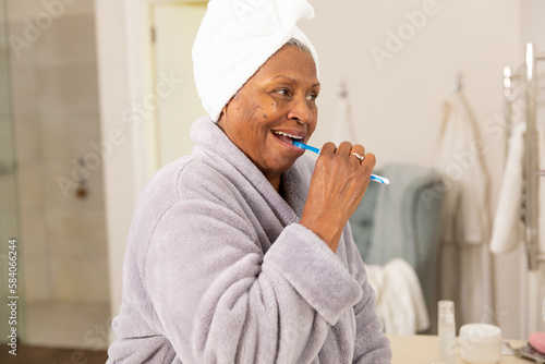 Side view of african american senior woman wearing bathrobe brushing teeth in bathroom photo