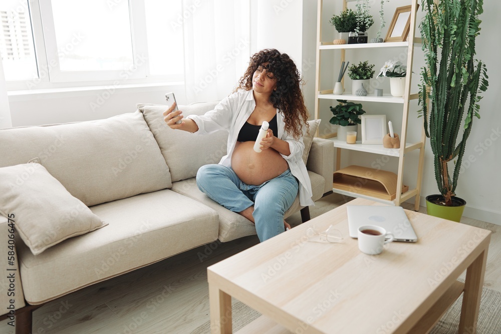 Pregnant woman smile blogger showing baby feeding bottle filming herself on phone, video chat sitting on sofa at home freelancer in last month of pregnancy lifestyle before childbirth Pregnant woman smile blogger showing baby feeding bottle filming herself on phone, video chat sitting on sofa at home freelancer in last month of pregnancy lifestyle before childbirth