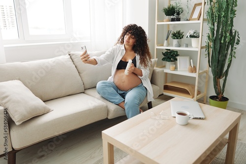 Pregnant woman smile blogger showing baby feeding bottle filming herself on phone, video chat sitting on sofa at home freelancer in last month of pregnancy lifestyle before childbirth