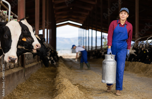 Portrait of young European woman dairy farmworker in uniform carrying metal milk can in cowshed