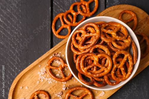 Board with bowl of tasty salted pretzels on dark wooden background, closeup photo