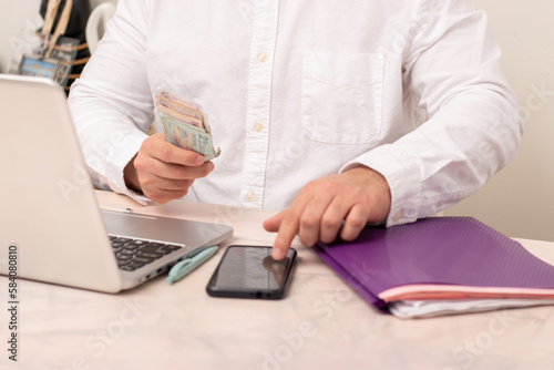 Horizontal view of Hispanic professional executive making a balance and counting money front their laptop in home office using calculate in their phone