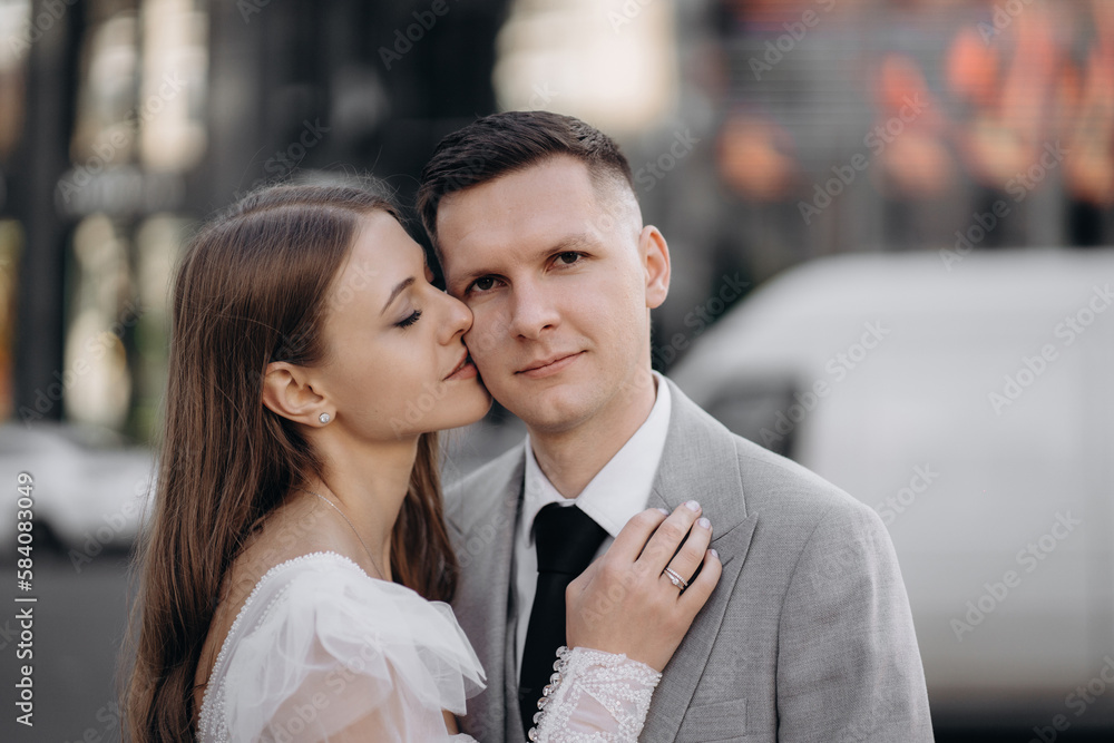 Front view of bride and groom getting married, having photoshoot outdoors. Woman wearing wedding dress, kissing groom, man looking at camera. Concept of marriage and love. Front view of bride and groom getting married, having photoshoot outdoors. Woman wearing wedding dress, kissing groom, man looking at camera. Concept of marriage and love.
