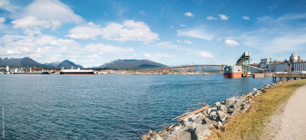 Cargo ship docked at port terminal in front of bridge and anchored container ships. Marine transport panorama with port facility. Iron workers Memorial Bridge, Burrard Inlet, Vancouver, BC, Canada.