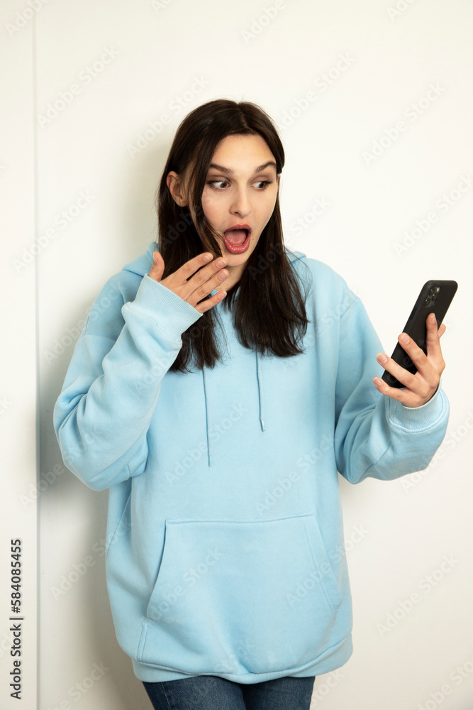 emotional girl with a phone in a blue jacket on a light background. The model is shocked. emotional girl with a phone in a blue jacket on a light background. The model is shocked.