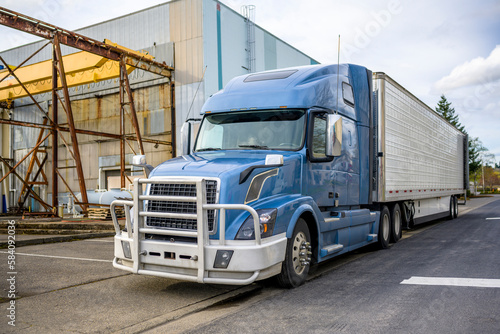Blue gray big rig semi truck with aluminum grille guard and refrigerator semi trailer with open back door standing near the warehouse on the street at industrial area waiting for the next load