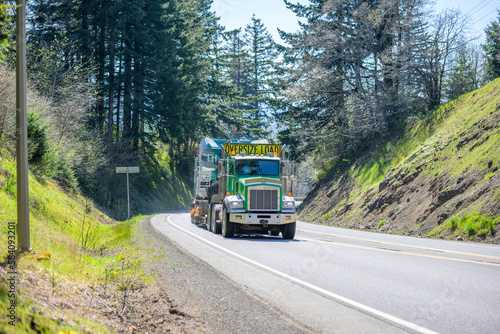 Classic American bonnet green big rig semi truck with oversize load sign on the roof transporting oversized equipment on step down semi trailer driving on the winding narrow mountain road