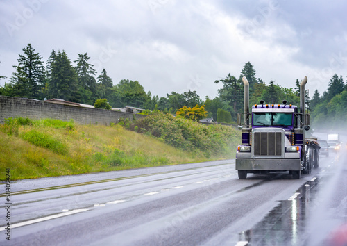 Day cab classic big rig American semi truck with empty flat bed semi trailer driving on the wet slippery highway road at raining weather