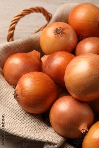 Wicker basket with many ripe onions on wooden table, closeup