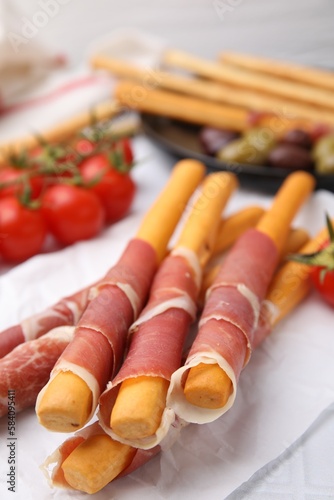 Delicious grissini sticks with prosciutto and snacks on white table, closeup