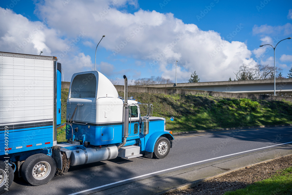Two colors classic big rig semi truck with roof spoiler transporting frozen cargo in refrigerated semi trailer driving on the highway road entrance at sunny day