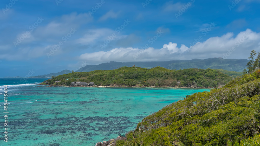 Corals are visible through the clear water of the turquoise ocean. The slopes of the islands are overgrown with lush tropical vegetation. In the distance, the villas of the hotel are visible.Seychelle