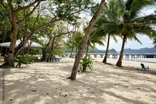 White sandy beach overgrown with palm trees at El Nideo, Palawan in the Philippines, with a jetty leading into the water.